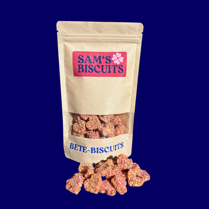 Tüte Bete-Biscuits. Schräg fotografierte hundekekse mit Gemüse von SAM'S BISCUITS. Vor der Tüte auf blauem Hintergrund liegen besondere leckerlis für Hunde in Herzform.
