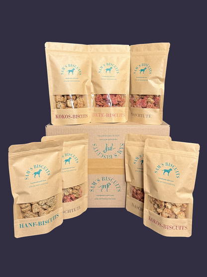 Sieben Tüten voller besonderer Hundekekse vegan vor einem Versandkarton von SAM'S BISCUITS